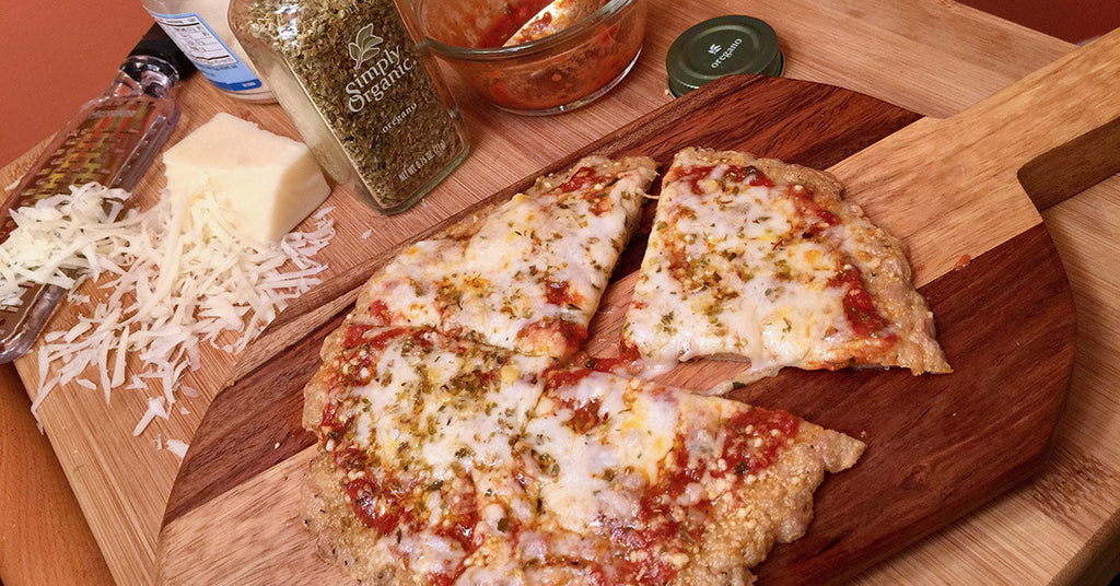 Homemade cheese pizza on a wooden board with grated cheese, oregano, and sauce ingredients nearby.