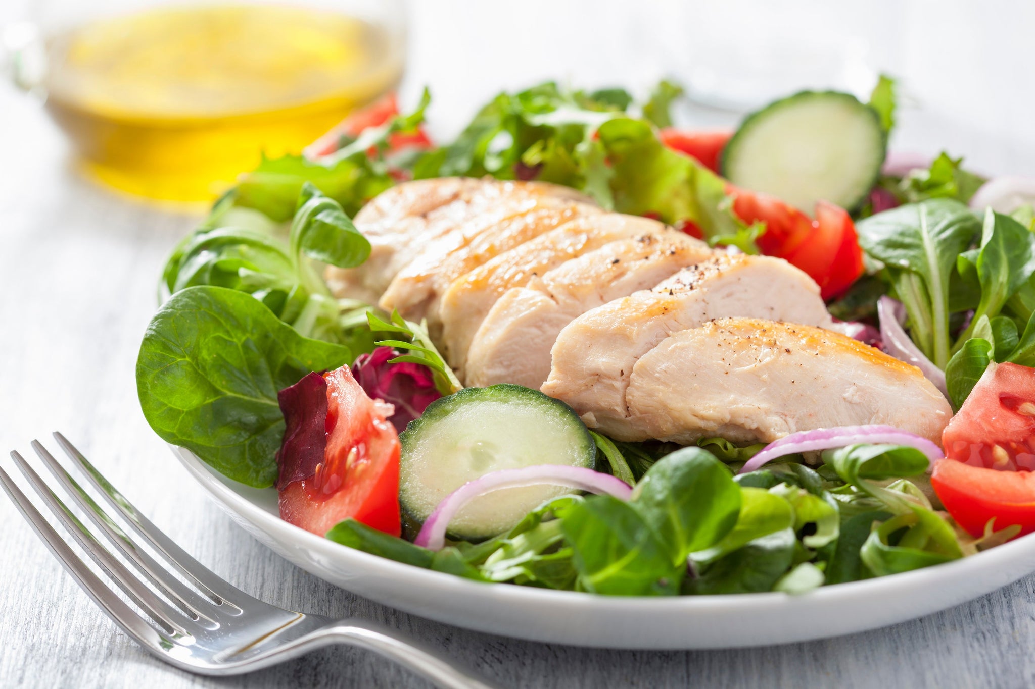 Fresh salad with sliced grilled chicken, tomatoes, cucumbers, greens, and red onion on a white plate.