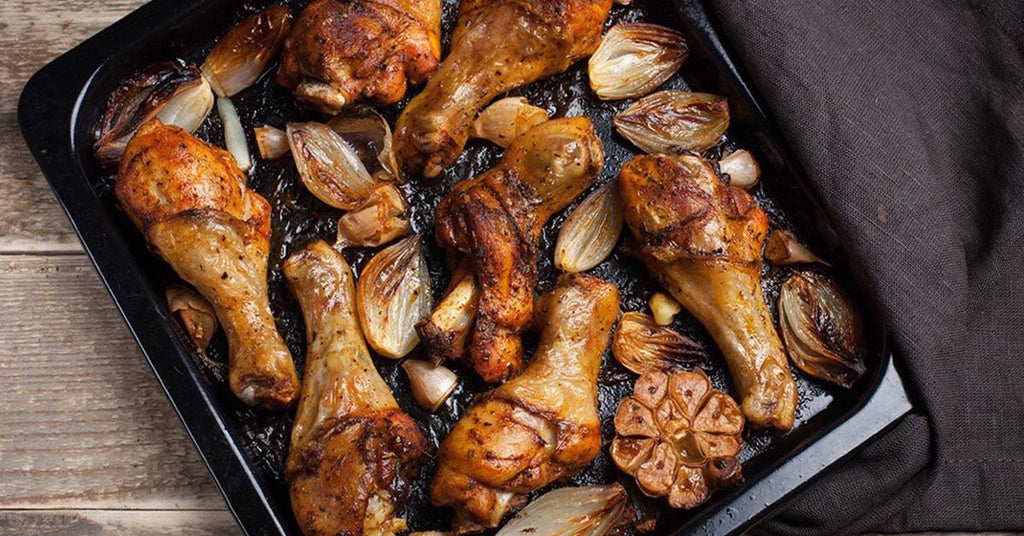 Roasted chicken drumsticks with caramelized onions and garlic on a dark baking tray.