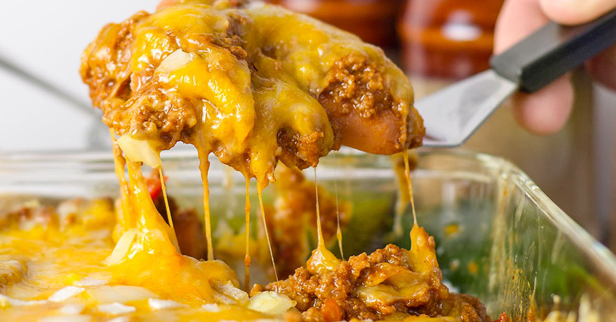 A spatula lifts a cheesy, baked dish with ground meat and sauce, showcasing gooey melted cheese stretching from the casserole in a kitchen setting.
