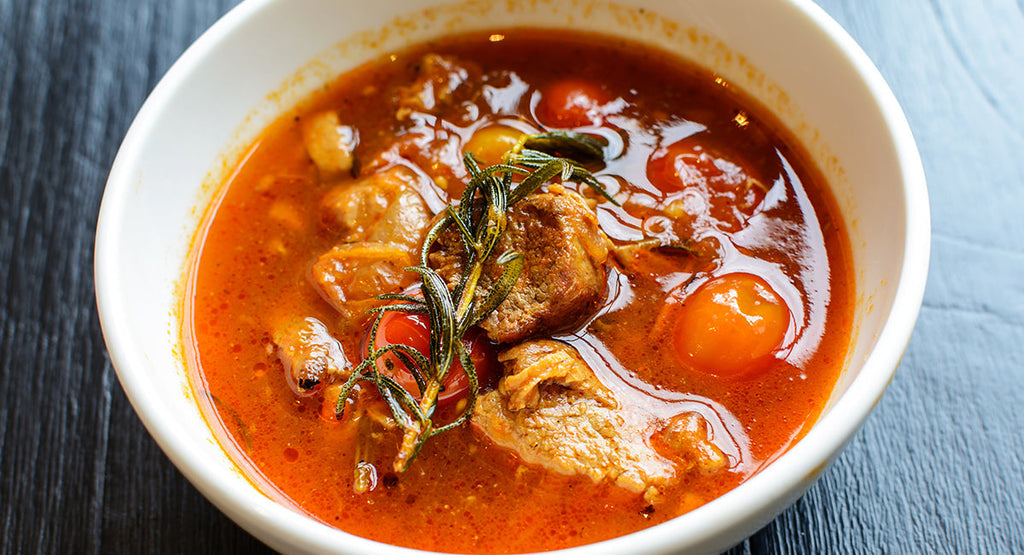 Bowl of rich tomato-based stew with chunks of meat, cherry tomatoes, and a sprig of rosemary.