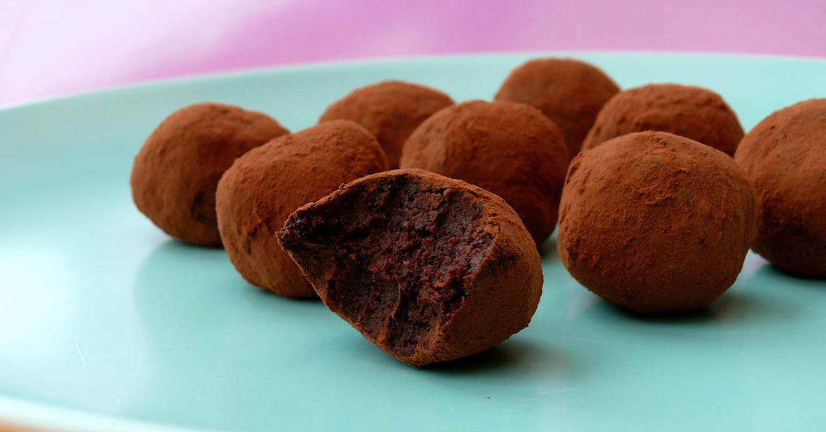 A close-up of several chocolate truffles coated in cocoa powder on a light blue plate, with one truffle partially bitten to reveal its rich, dark interior.