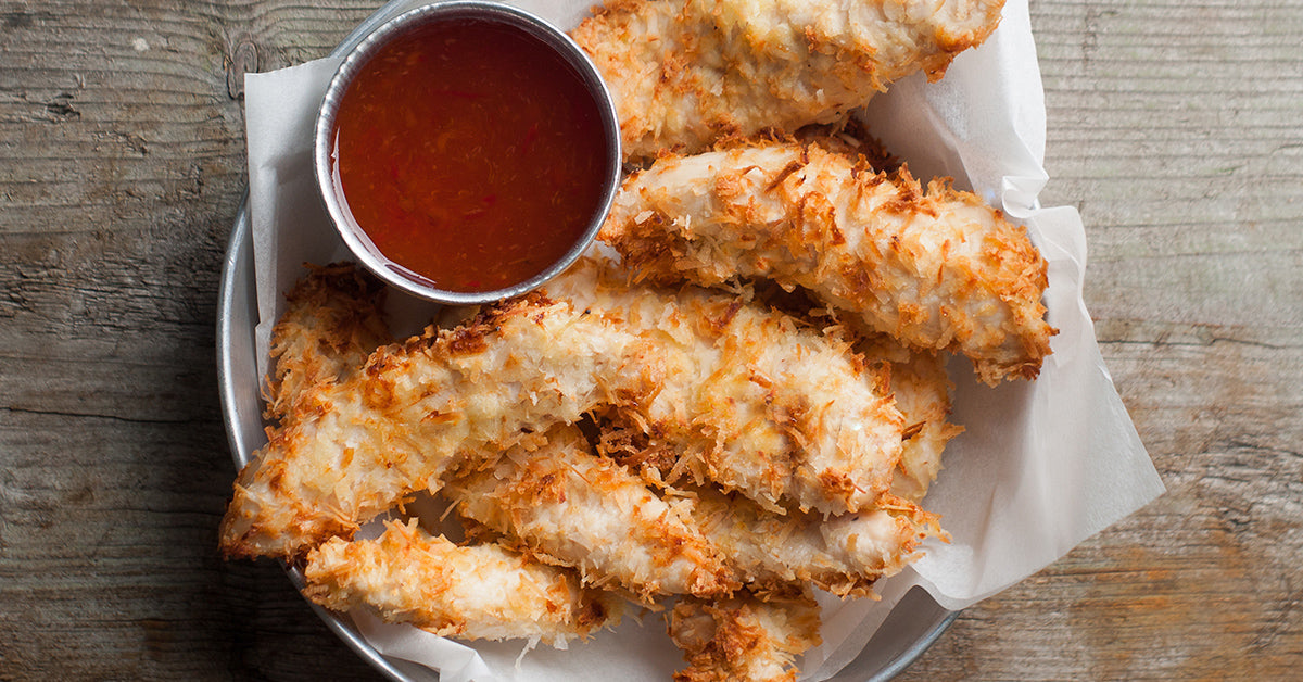 Crispy coconut-crusted chicken tenders served in a basket with a small cup of sweet chili dipping sauce.