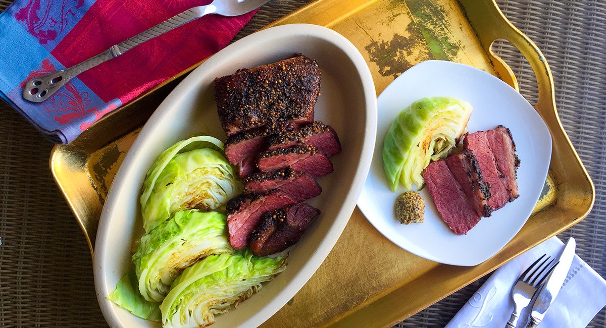 Sliced corned beef served with wedges of cooked cabbage and mustard on a gold tray with utensils and napkins.