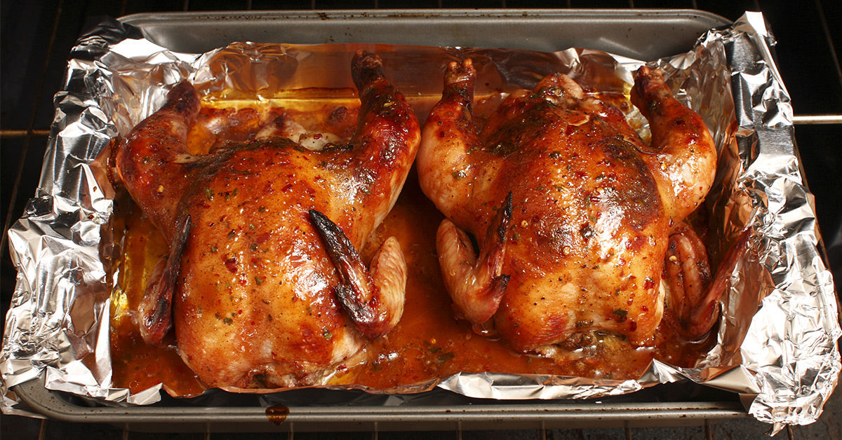 Two roasted chickens are placed side by side in a metal baking pan lined with foil, glistening with a savory glaze and surrounded by juices, inside an oven.