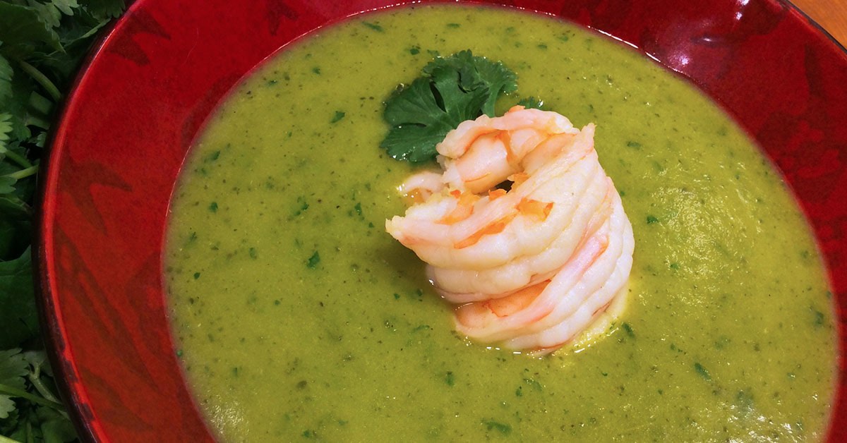 Green creamy soup topped with cooked shrimp and a cilantro leaf in a red bowl.