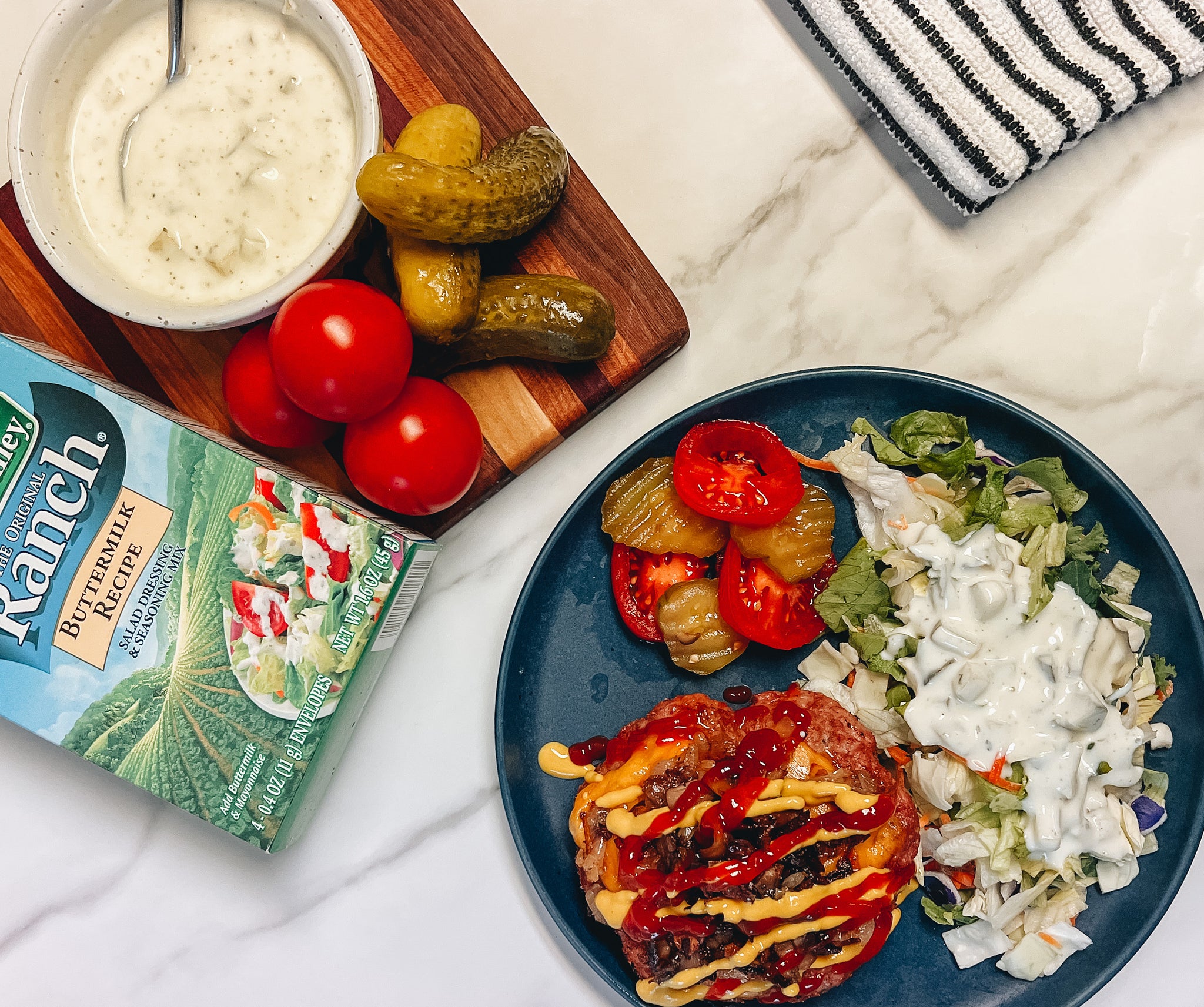 A plate features a burger topped with ketchup and mustard, alongside a salad with ranch dressing, pickles, and sliced tomatoes, with a bowl of ranch dressing and fresh tomatoes on a wooden cutting board nearby.