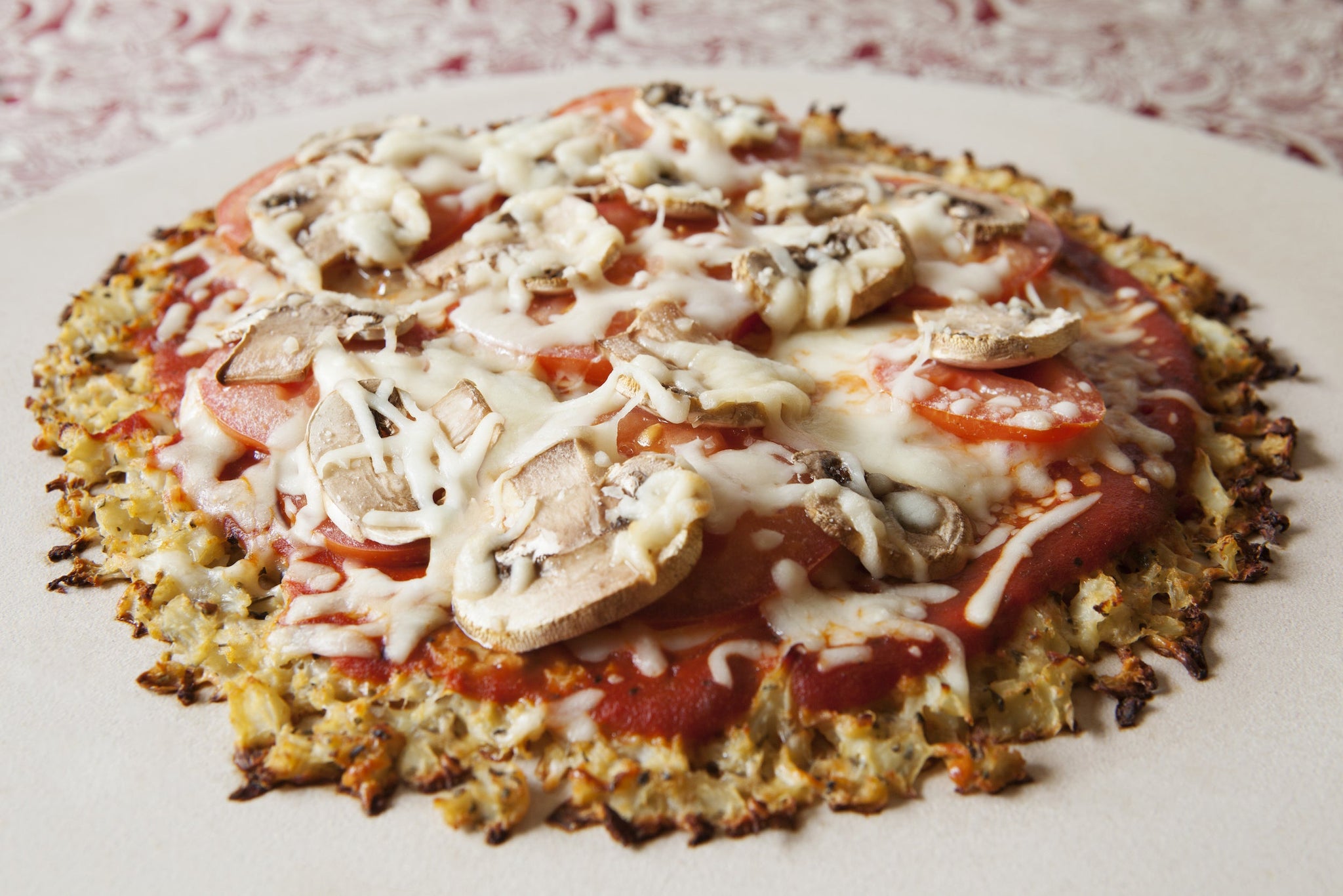 Cauliflower-crust pizza topped with tomato sauce, sliced tomatoes, mushrooms, and melted cheese on a light surface.