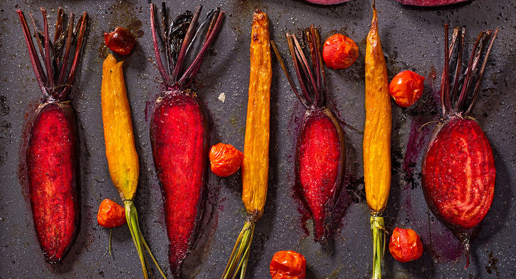 A variety of colorful roasted beets and carrots are arranged on a dark baking sheet, with vibrant red and orange hues and some cherry tomatoes scattered among them.