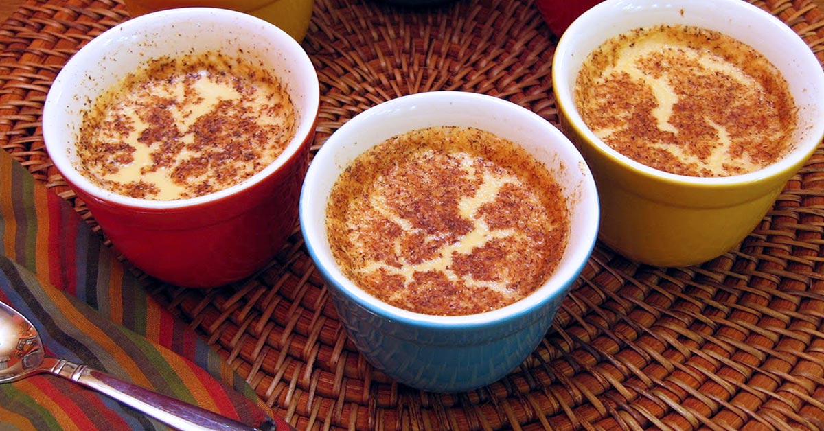 Creamy custard desserts in colorful cups, topped with a dusting of cinnamon on a woven placemat.