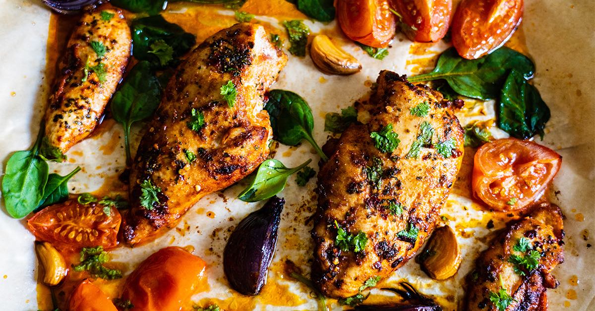 Grilled chicken breasts seasoned with herbs are arranged on parchment paper alongside roasted tomatoes, garlic, and fresh spinach, all glistening with flavorful juices.