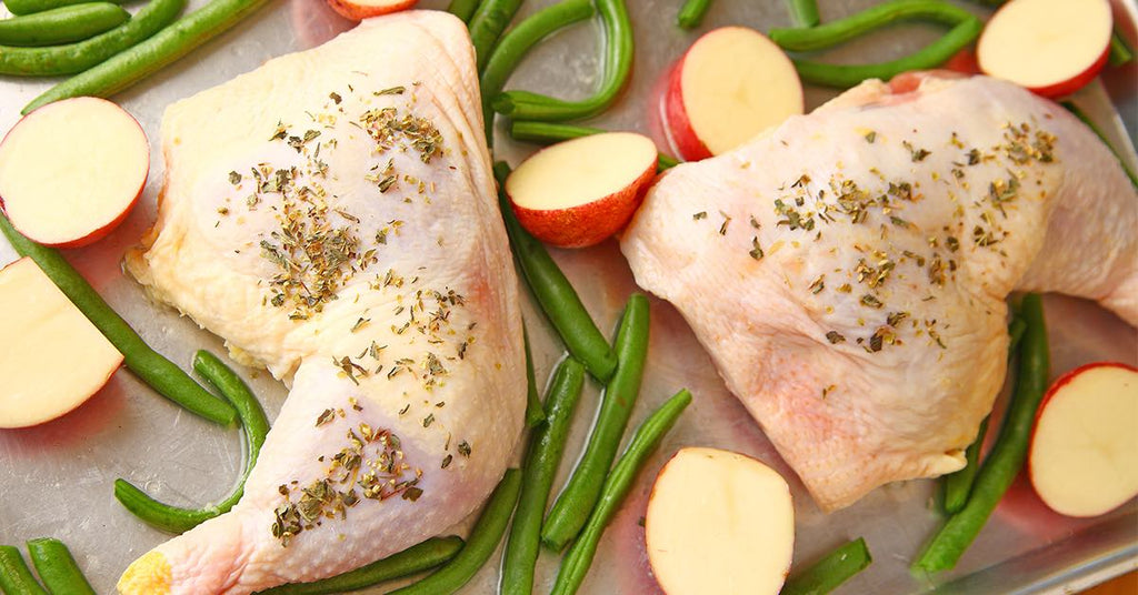 Raw chicken legs seasoned with herbs on a tray with green beans and halved red potatoes.