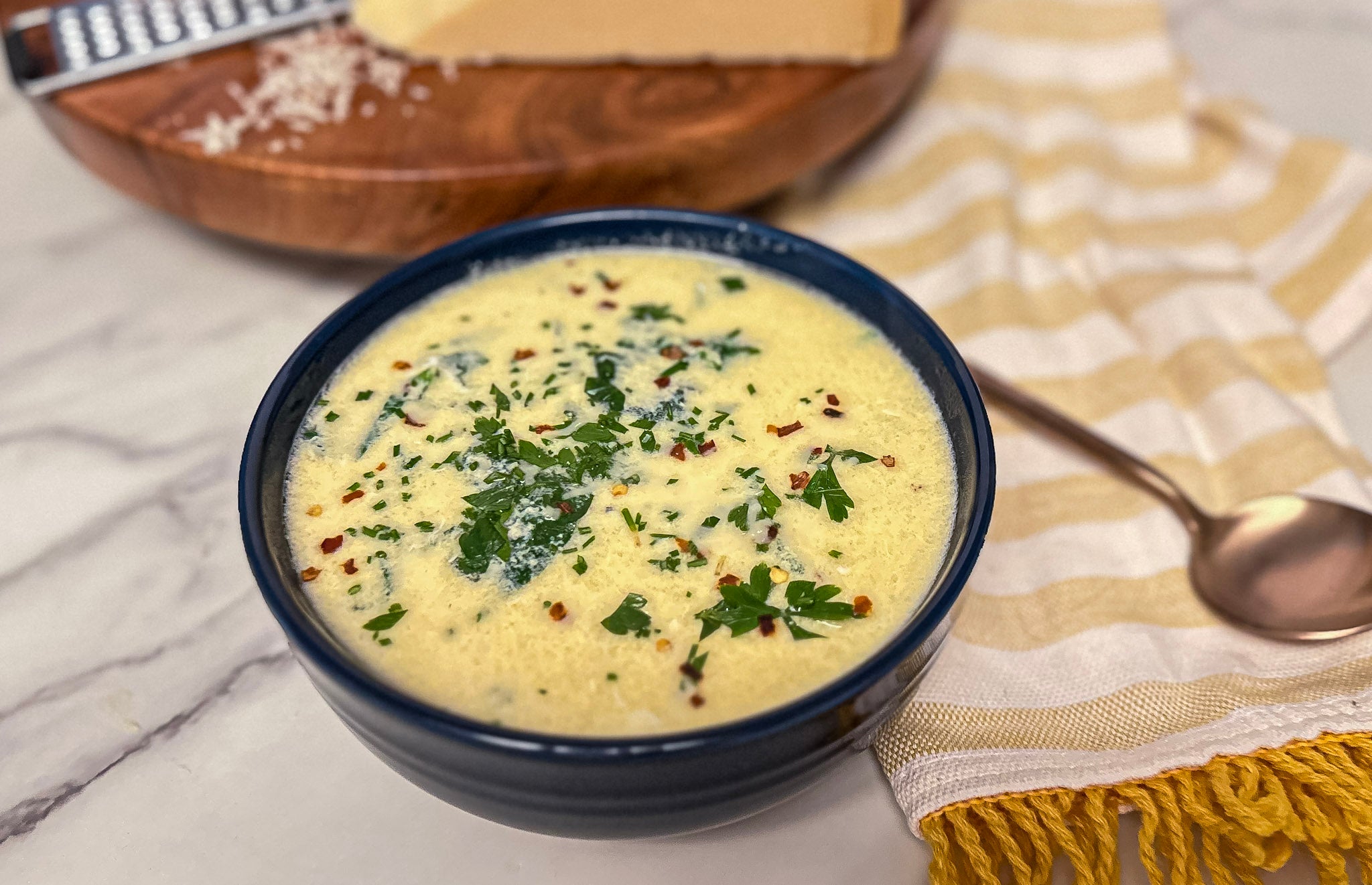 A blue bowl filled with creamy soup topped with parsley and red pepper flakes sits on a marble surface next to a striped towel and a wooden board with grated cheese.