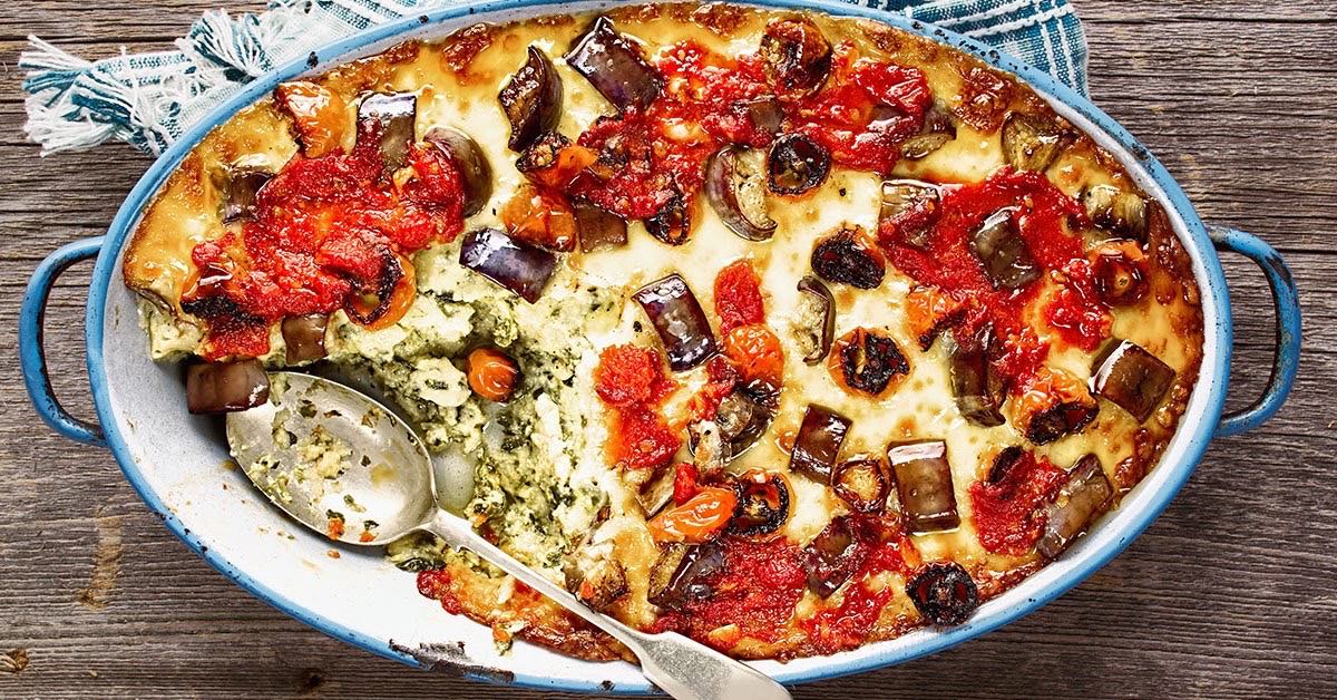 Baked casserole with eggplant, tomatoes, and cheese in a blue dish, partially scooped with a spoon.