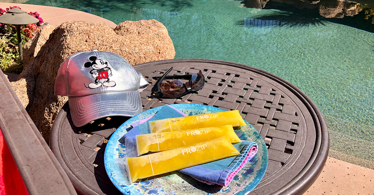 Plate with yellow freezer pops beside a silver Mickey Mouse hat and sunglasses on a poolside table.