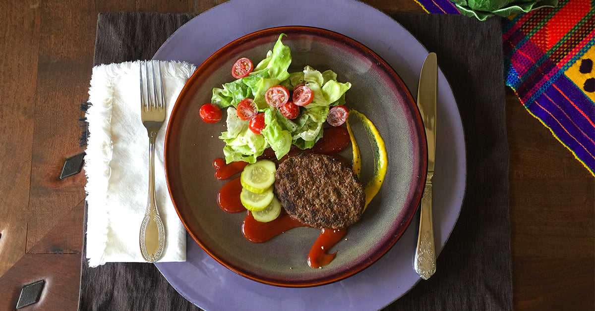 Plate with a burger patty, salad, cucumber slices, and sauces, set on a table with fork and knife.