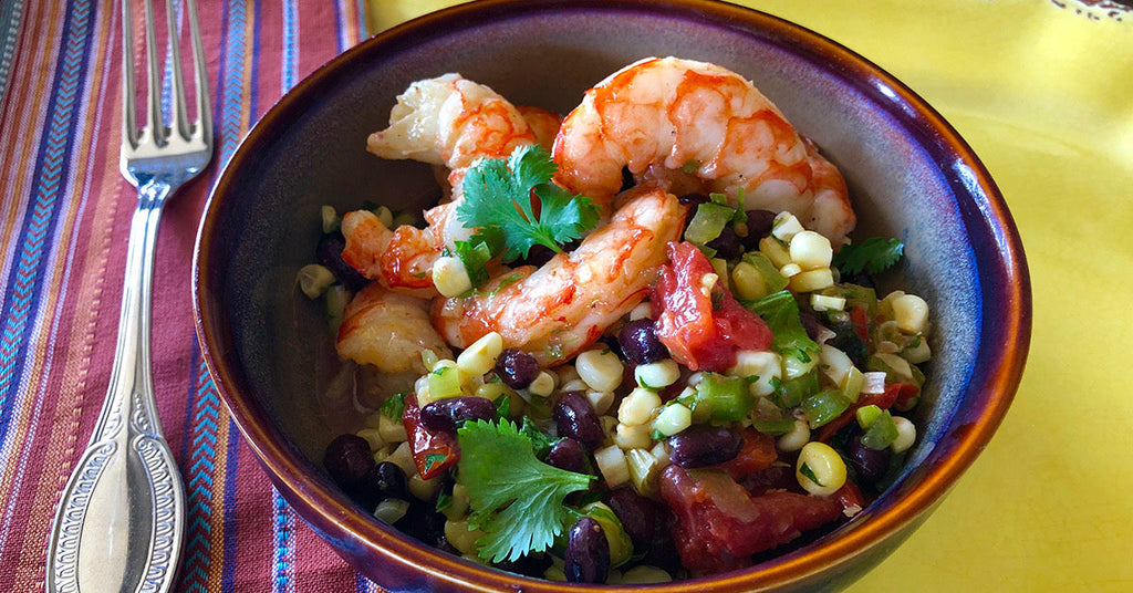 Bowl of shrimp with black beans, corn, tomatoes, and cilantro on a colorful table setting.