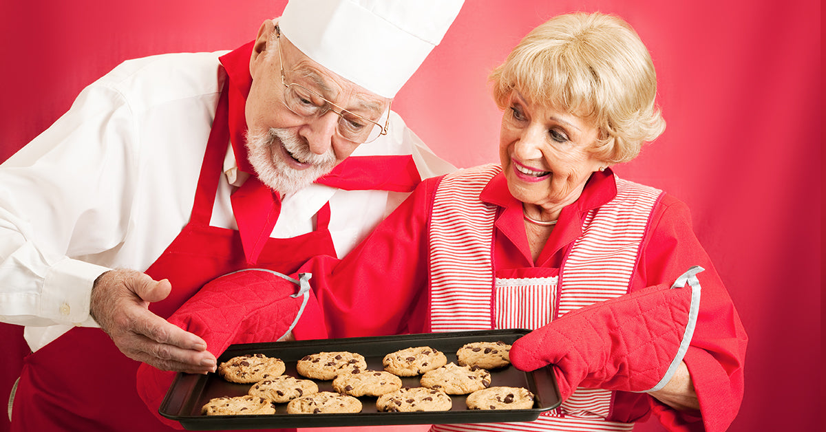 Two smiling older adults in red aprons holding a tray of freshly baked chocolate chip cookies.