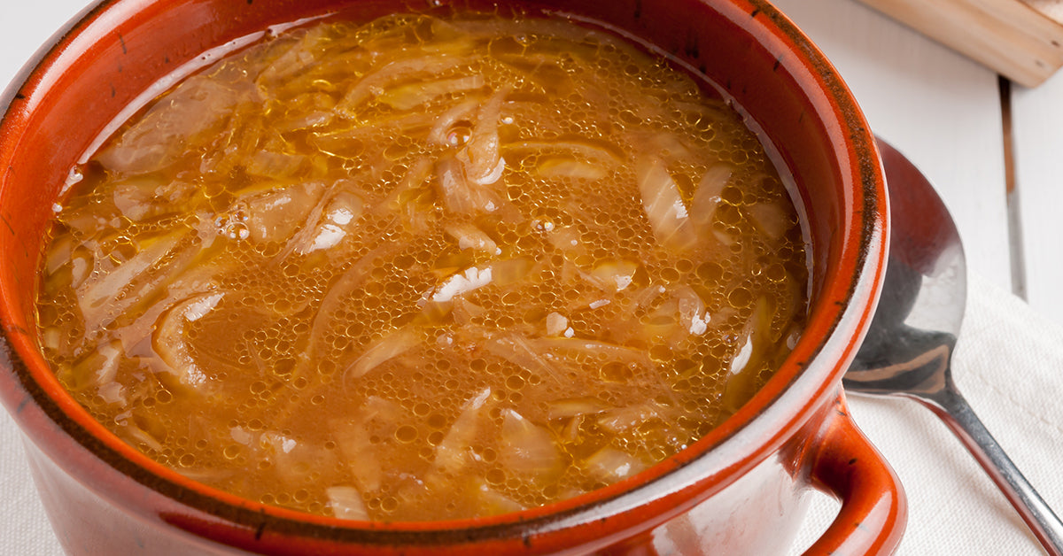 A red bowl filled with bubbling French onion soup, featuring thin slices of caramelized onions in a rich, golden broth, sits on a white tablecloth next to a silver spoon.