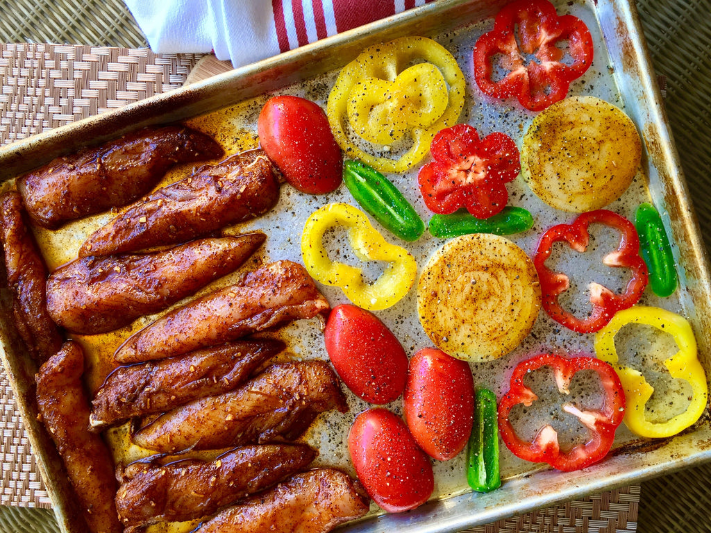 A baking tray displays seasoned chicken fillets alongside colorful sliced vegetables, including red and yellow bell peppers, tomatoes, and green peppers, ready for roasting.