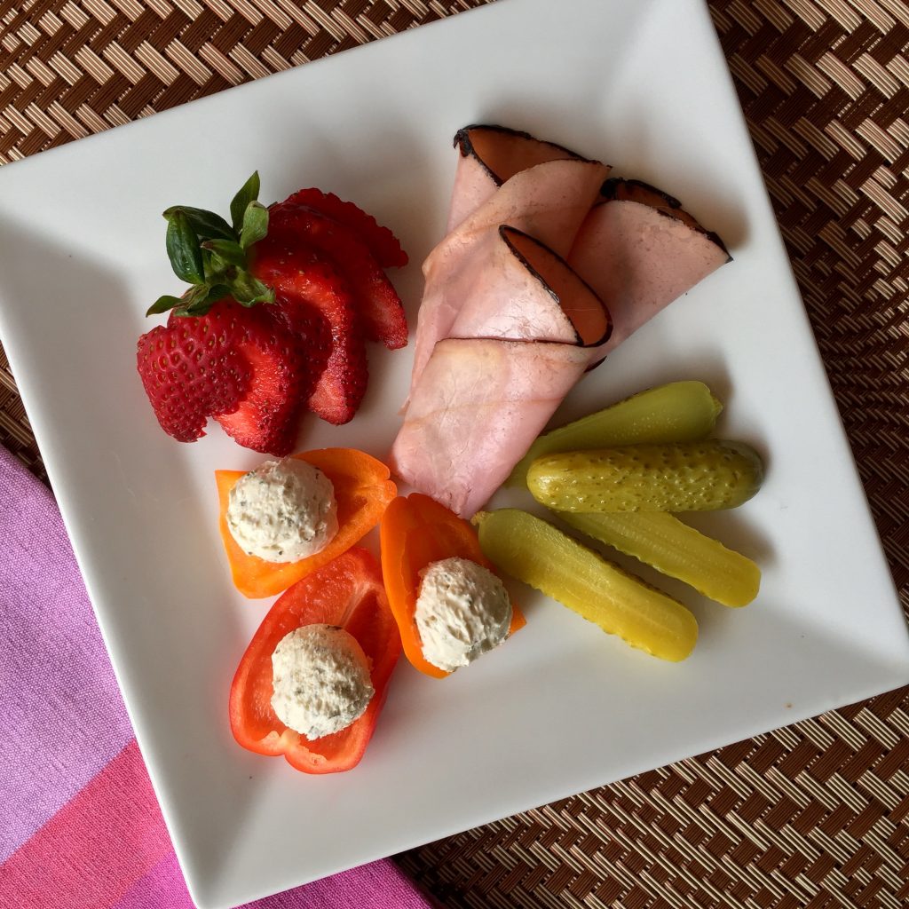 A square white plate with sliced strawberries, rolled slices of deli ham, small pickles, and bell pepper pieces topped with dollops of a creamy spread.