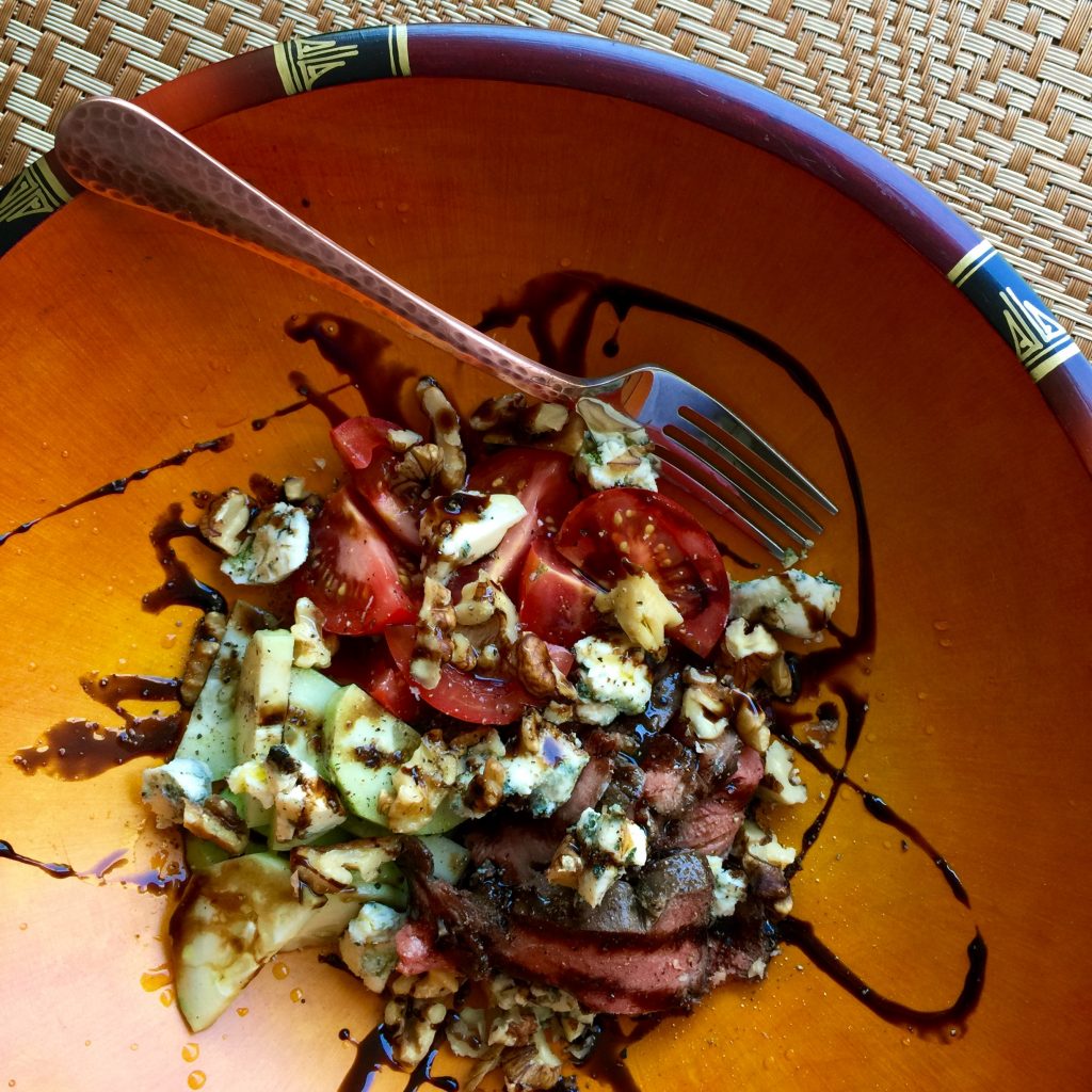 Salad with tomatoes, cucumbers, steak slices, walnuts, blue cheese, and balsamic drizzle in an orange bowl.