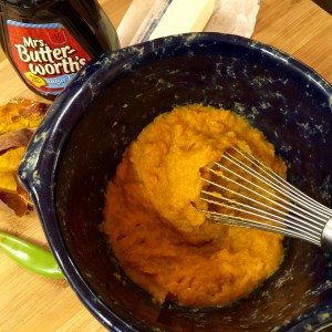 Whisk mixing orange mashed mixture in a bowl, with syrup bottle, butter, and cooked squash pieces nearby.