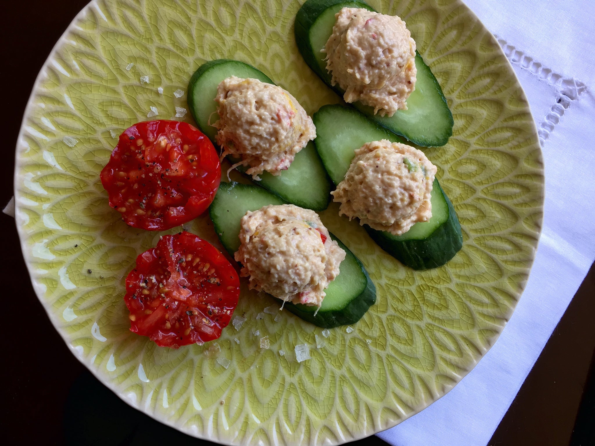A green plate holds cucumber slices topped with crab mixture and garnished with two slices of red tomato, all presented on a textured background.