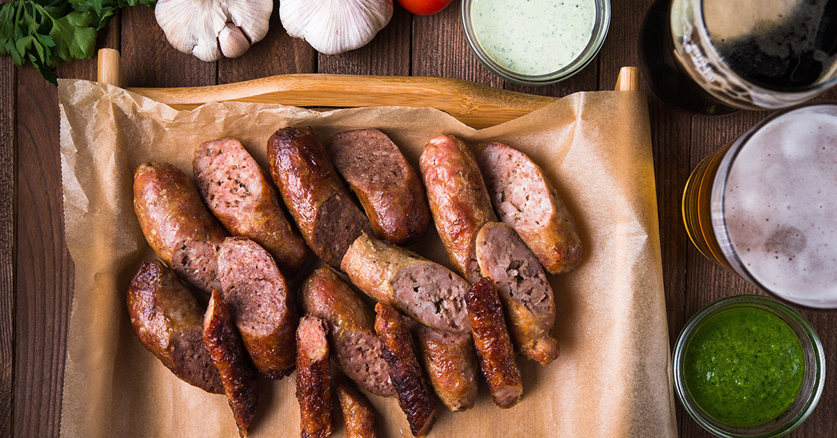 Grilled sliced sausages on parchment paper with dipping sauces and drinks on a wooden table.
