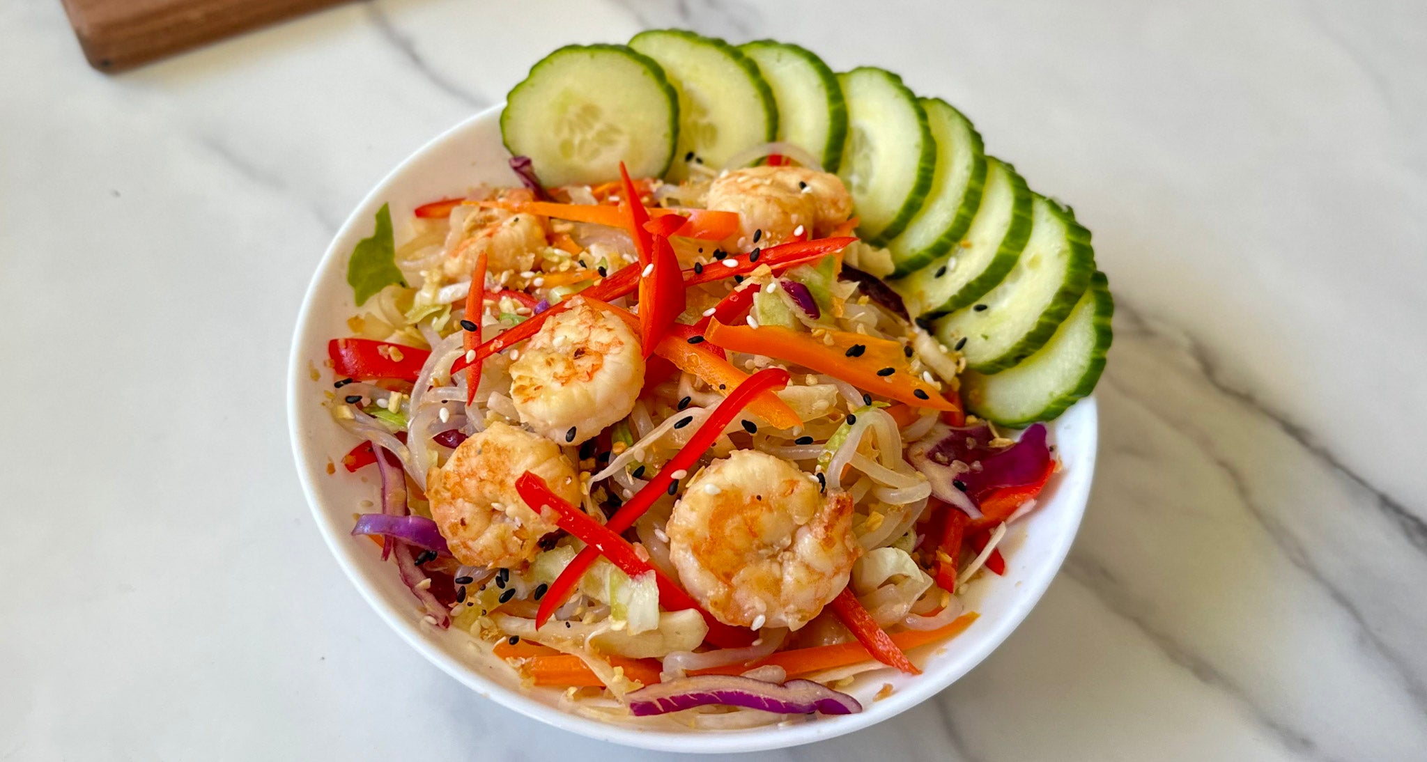 A white bowl filled with colorful shrimp salad features shrimp, sliced red and orange peppers, shredded cabbage, and cucumber, garnished with sesame seeds, on a marble countertop.