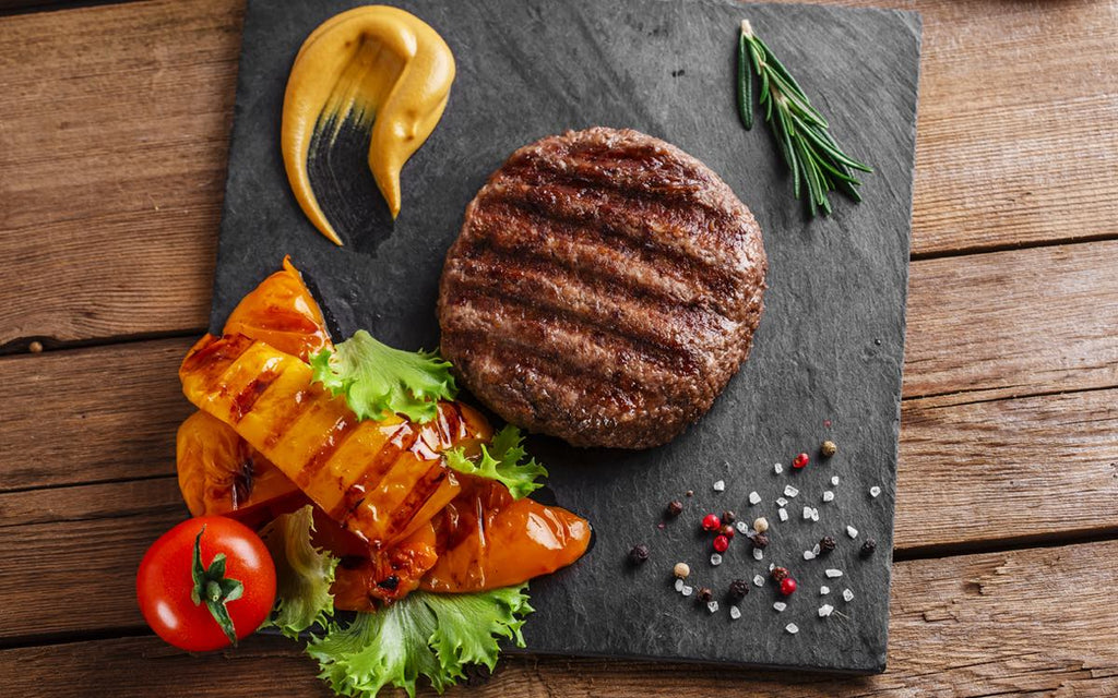 Grilled burger patty on a slate with roasted peppers, lettuce, tomato, mustard, rosemary, and scattered spices.