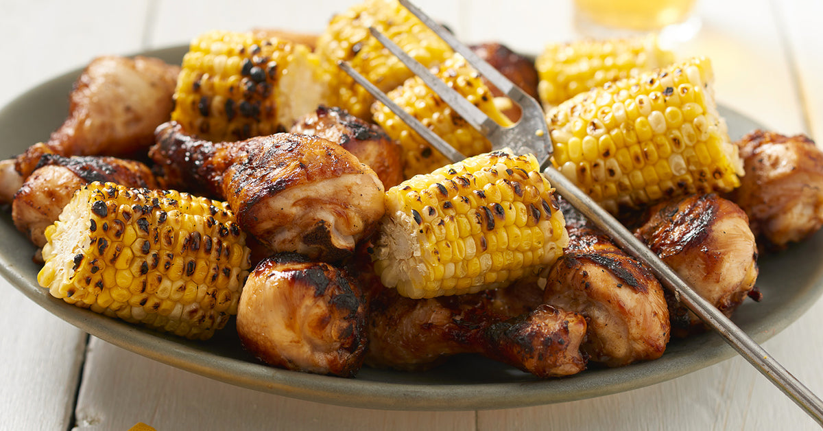 Grilled adobo seasoned chicken pieces and charred corn on the cob are arranged on a round plate, with a fork resting on top, set against a light wooden background.