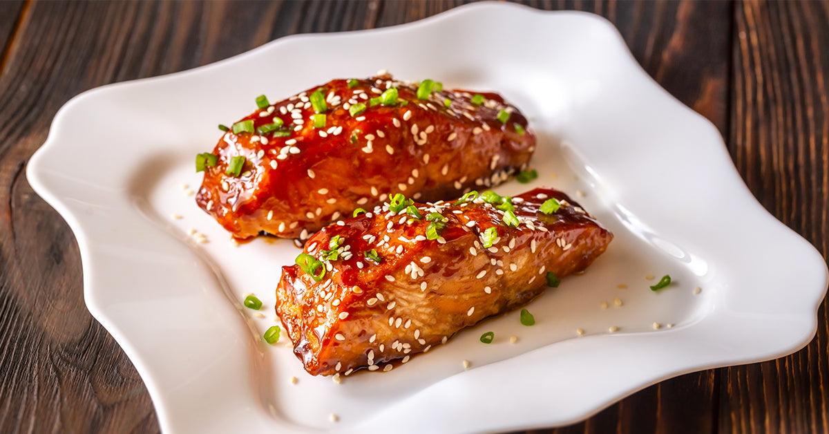 Two glazed meatloaf pieces topped with chopped green onions and sesame seeds are served on a decorative white plate, set against a rustic wooden background.