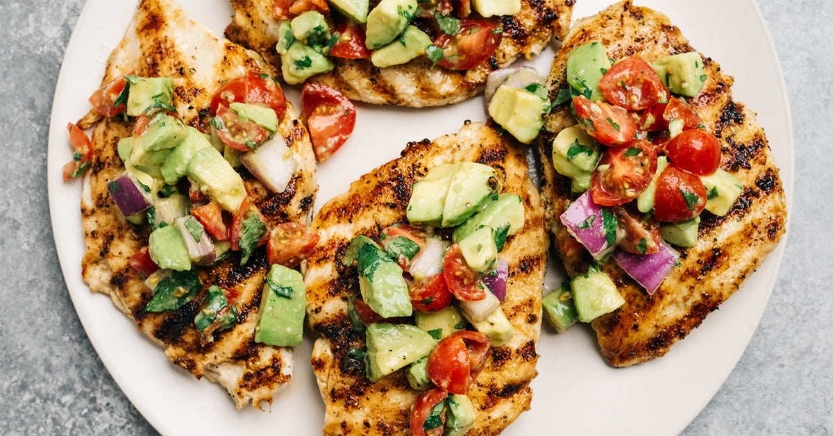 Grilled chicken fillets are topped with a colorful mixture of diced tomatoes, avocado, and herbs, arranged neatly on a round white plate against a light gray background.