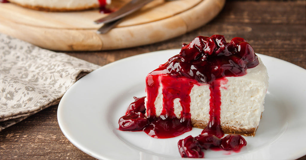 Slice of cheesecake on a white plate topped with glossy cherry sauce dripping down the sides.