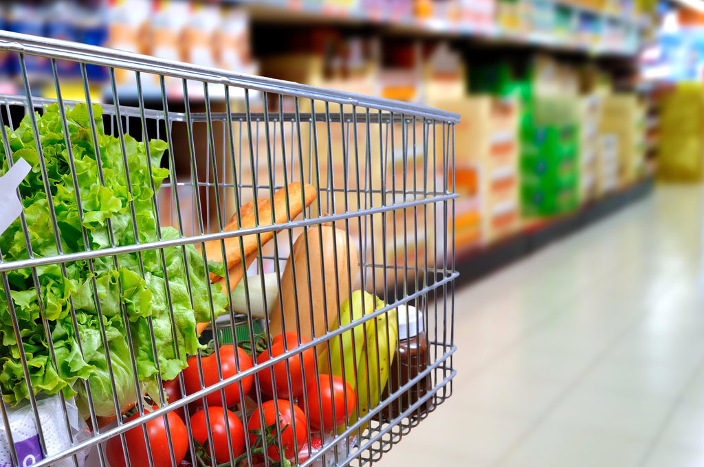 A shopping cart filled with fresh produce, including lettuce, tomatoes, bananas, and bread, is positioned in a grocery store aisle with colorful product displays in the background.