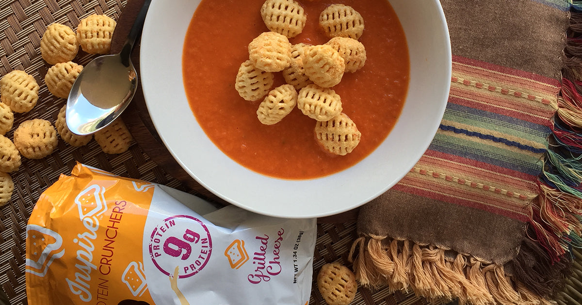A bowl of orange soup topped with crunchy, waffle-shaped snacks sits on a woven mat, accompanied by a spoon and a partially visible bag of protein crunchers labeled "Grilled Cheese."