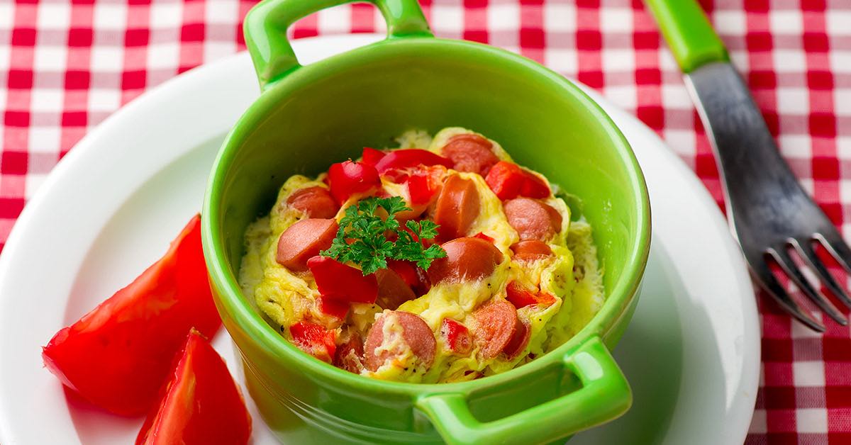 A green bowl holds a hot dog omelet topped with diced tomatoes and parsley, accompanied by slices of red tomato, set on a red and white checkered tablecloth.