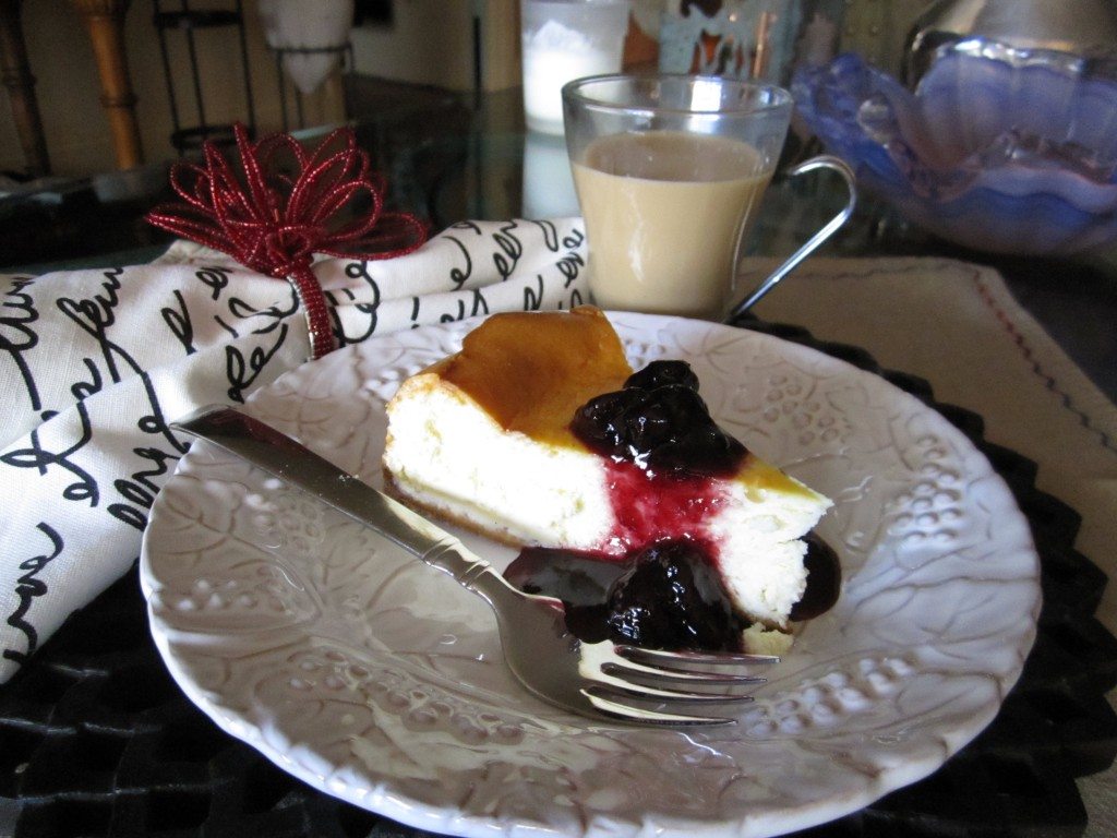 Slice of cheesecake with berry topping on a white plate beside a fork and a glass mug of coffee.