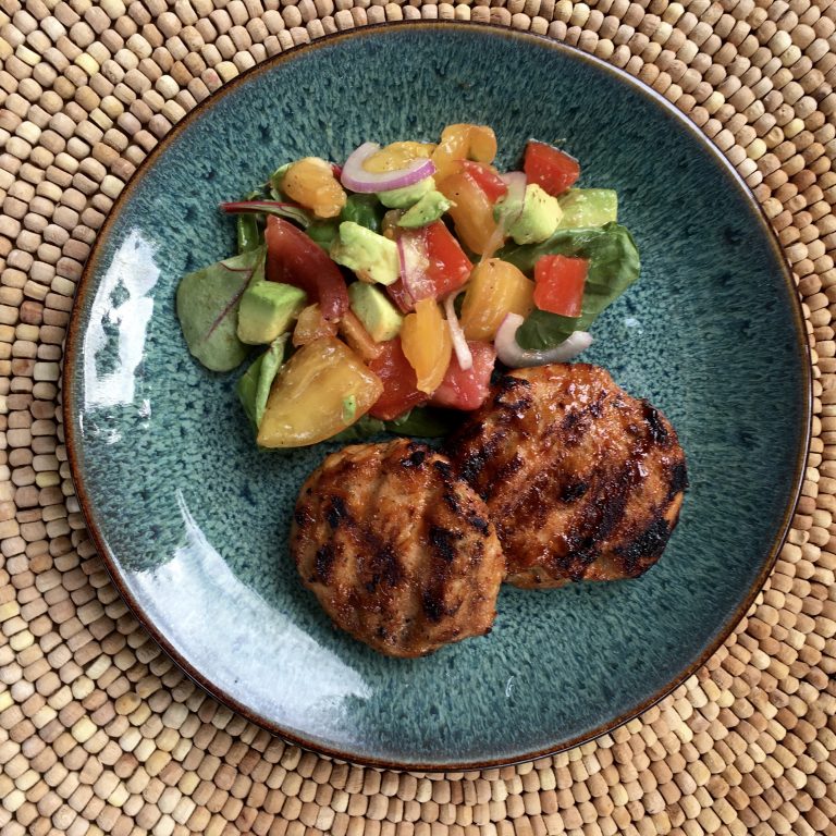 A green plate holds two grilled patties beside a colorful salad made of diced tomatoes, avocados, and onions, all set on a woven placemat.