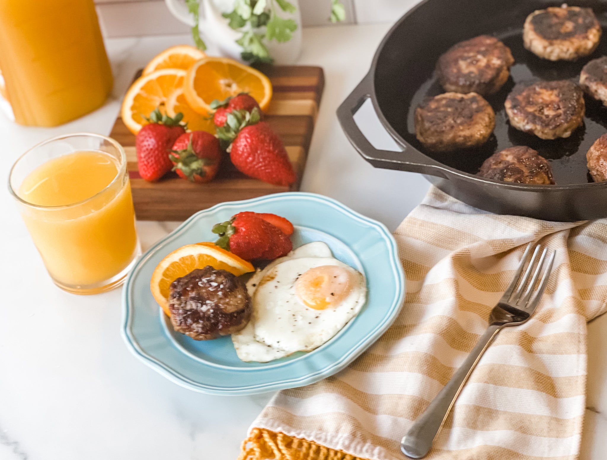 A blue plate holds a sausage patty, fried egg, and strawberries with an orange slice, alongside a glass of orange juice, while a cast iron skillet with more sausage patties sits nearby on a striped towel.