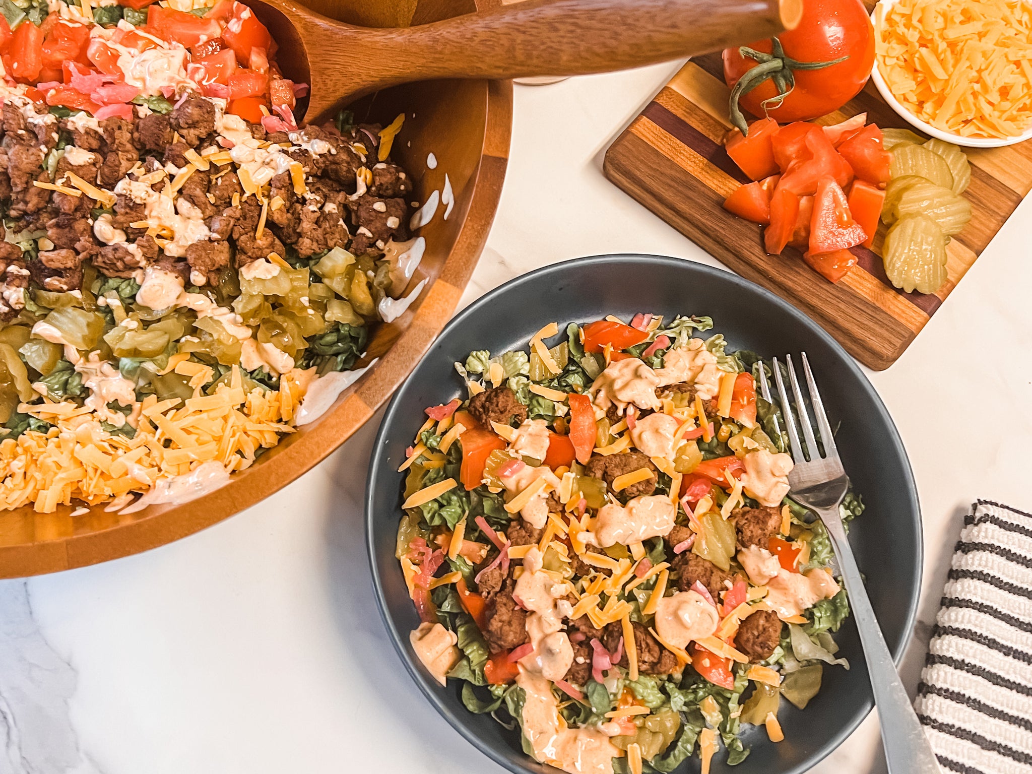 A bowl of Big Mac-inspired salad features layers of lettuce, ground beef, pickles, tomatoes, cheese, and special sauce, with a wooden spoon resting in a larger bowl and chopped ingredients on a cutting board nearby.