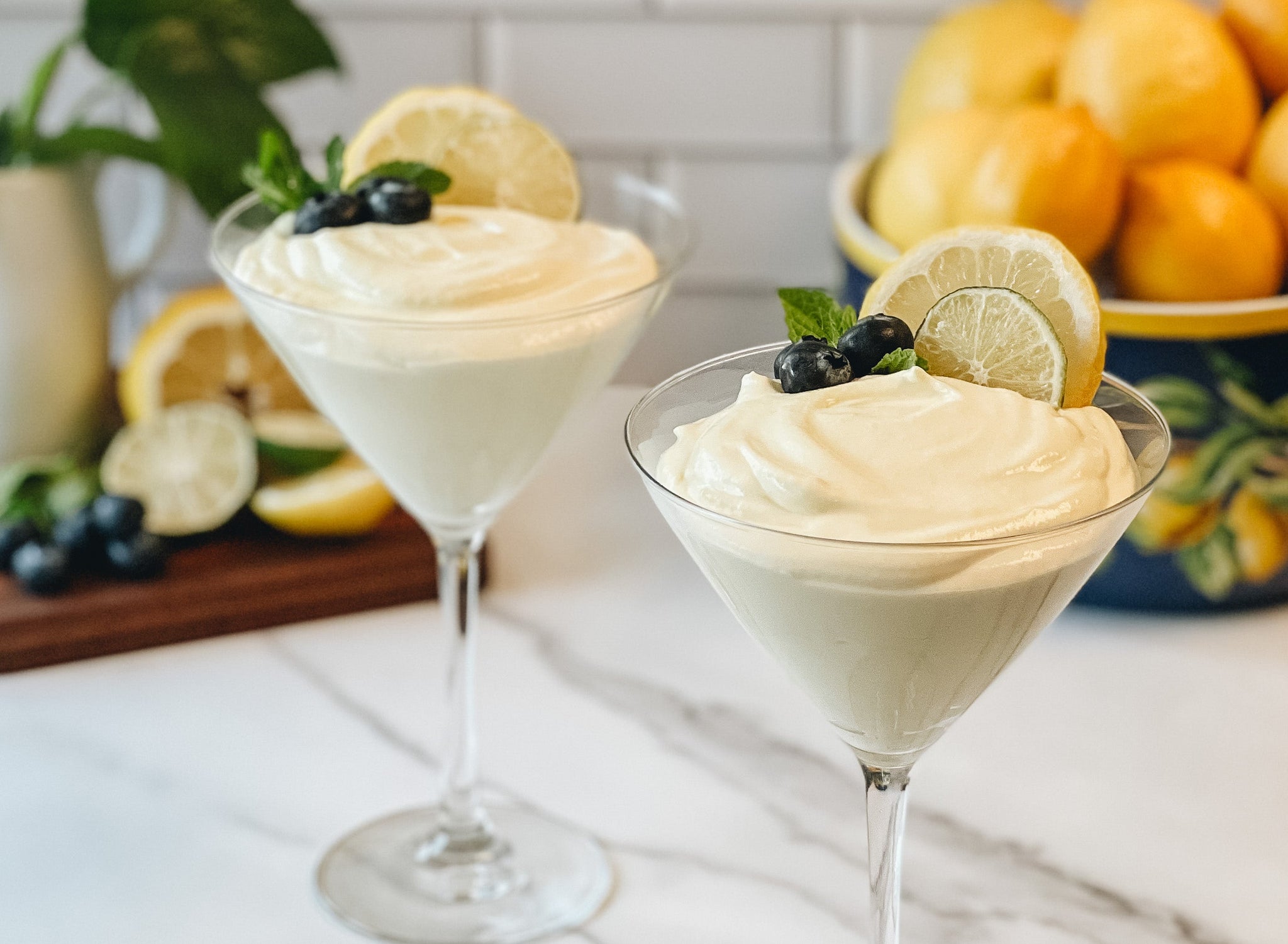 Two elegant dessert glasses filled with creamy lemon mousse are garnished with a slice of lemon, blueberries, and mint, set against a backdrop of fresh lemons and greenery.