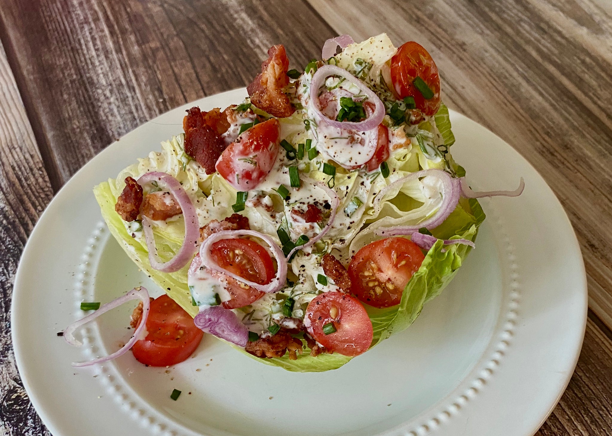 A wedge of lettuce topped with creamy dressing, crispy bacon, sliced tomatoes, red onions, and chives sits on a white plate against a wooden background.