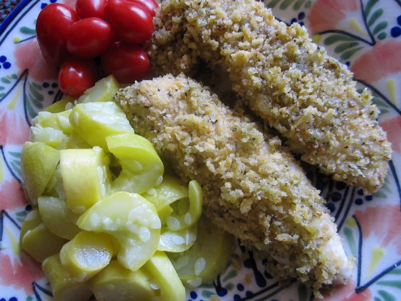 A colorful plate features two breaded fish fillets, a serving of sliced yellow squash, and a handful of cherry tomatoes, all arranged on a decorative patterned dish.