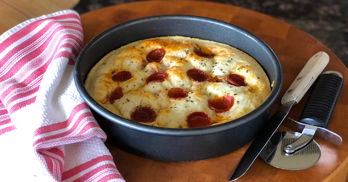 A round pan of freshly baked pizza is topped with pepperoni and herbs, resting on a wooden surface next to a striped kitchen towel and a pizza cutter.