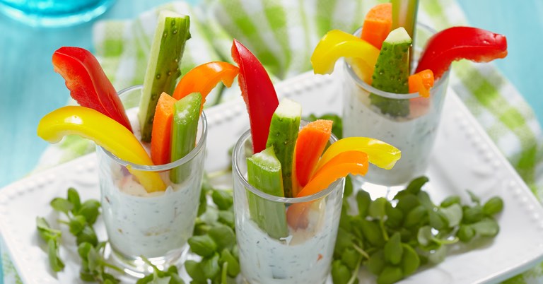 Colorful veggie sticks in small glasses of herb dip, including peppers, cucumbers, and carrots.