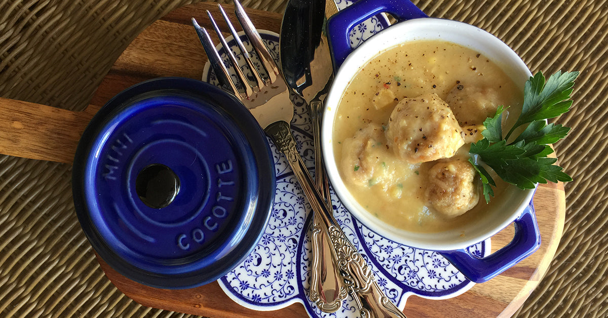 A small blue cocotte filled with chicken meatballs in creamy corn sauce sits on a decorative plate, accompanied by elegant silverware and garnished with a sprig of parsley.