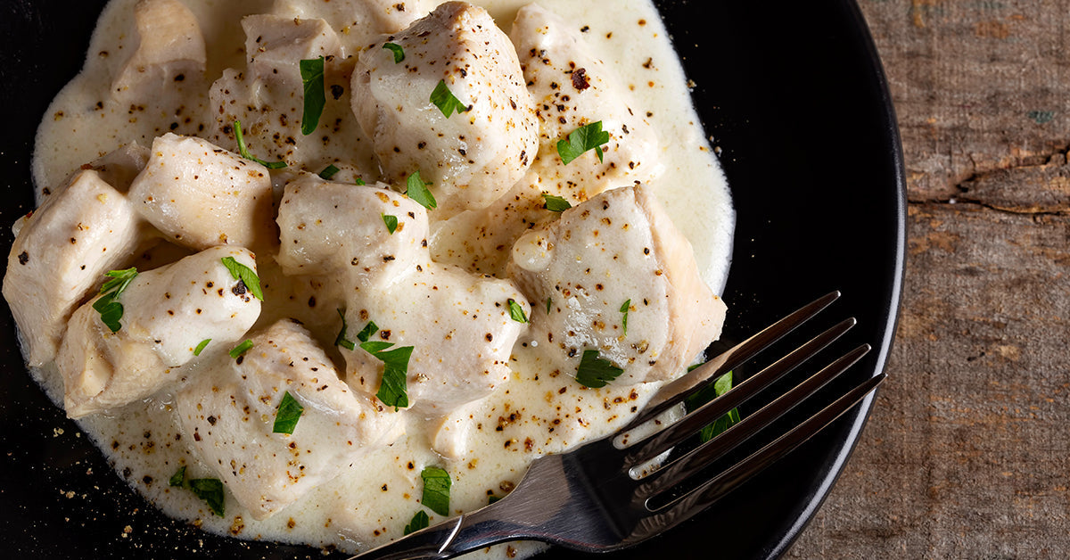 Tender pieces of chicken in a creamy mushroom sauce are garnished with fresh parsley on a black plate, accompanied by a silver fork, set against a rustic wooden background.