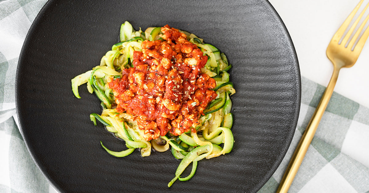 Zucchini spaghetti topped with a hearty tomato and meat sauce is served on a black plate, accompanied by a gold fork and a checkered napkin in the background.